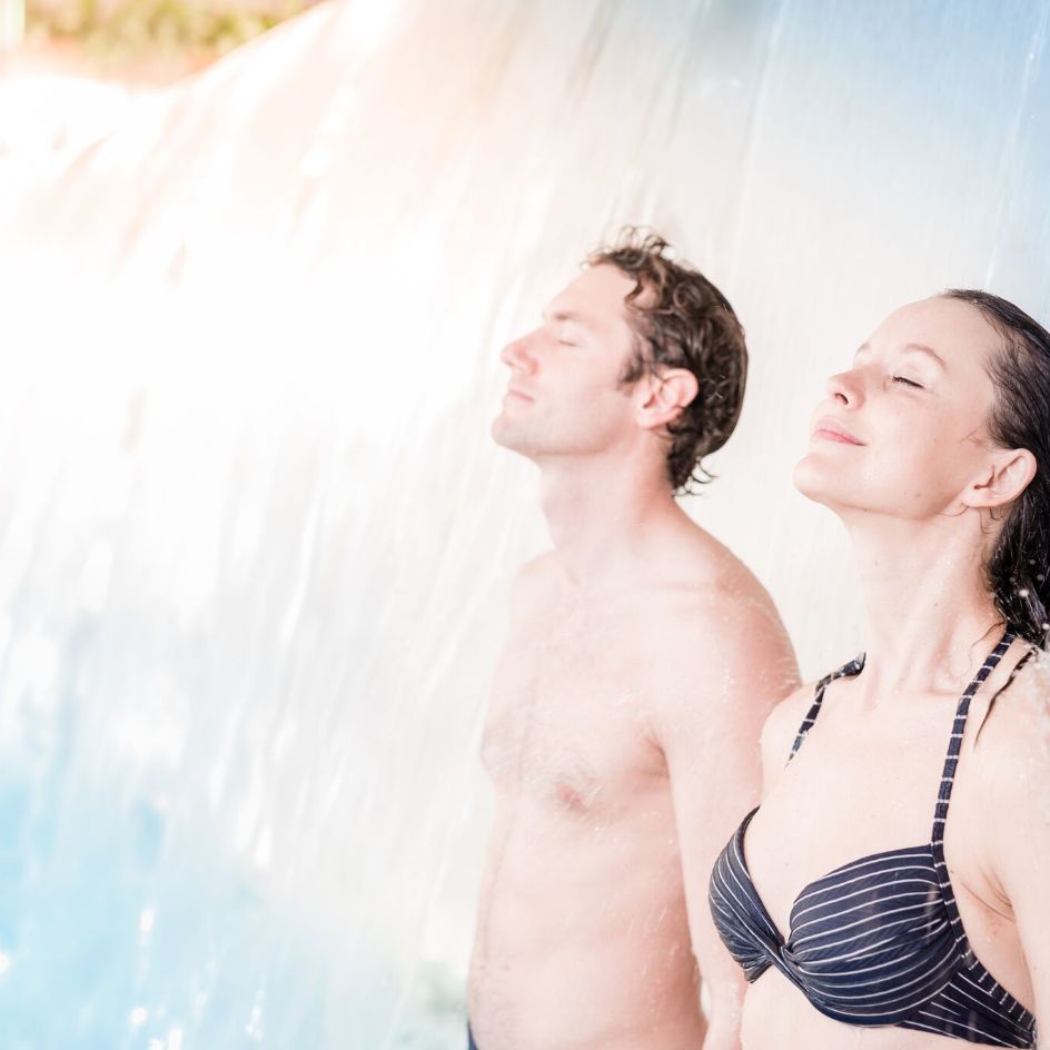 Pärchen steht unter dem Wasserfall der Therme NOVA und genießt das über den Rücken fließende Wasser.